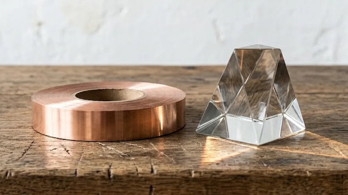 Prisme de verre transparent et rouleau de cuivre sur un établi en chêne brut éclairé par une lumière rasante.